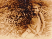 A sepia pic of a young boy in dungarees, sitting alone in what looks like hay.

