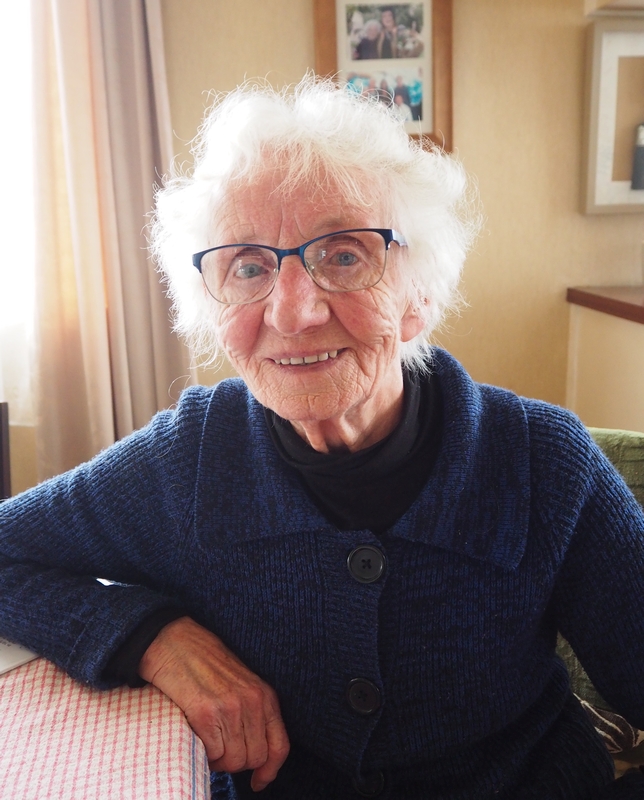 Another smiling old lady, this time sitting and leaning her elbow on a table. Royal blue top, same coloured glasses and still blue eyes. Shortish white hair.

