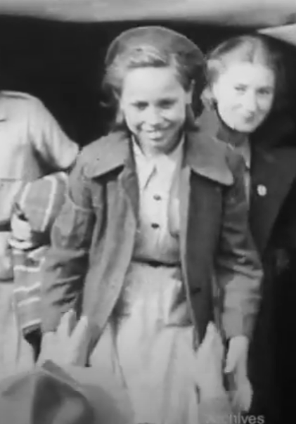 A smiling 13-year-old in a beret, dress and jacket looking towards hands outstretched to help her off the back of a truck. 