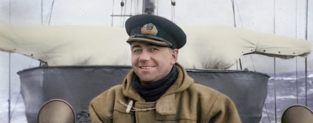 A duffel-coated commander probably on the bridge of his ship, smiling at the camera. Seas behind him are rough and grey.